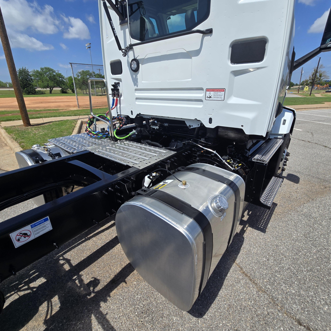Front-left side of a new Mack AN64T conventional daycab showing fuel tank and chassis with exposed wiring and steps.