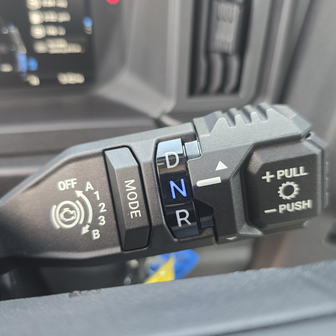 Truck gear selector with D, N, R markings and mode button in focus; adjacent push/pull control on steering column.
