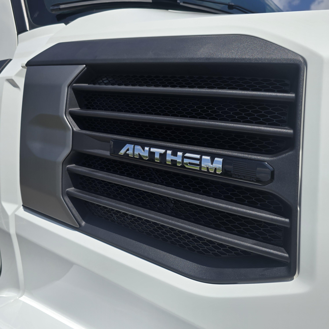 Front grille of a white Mack truck showing the ANTHEM badge in the center with black mesh and horizontal slats.