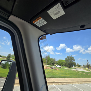 Cab interior view of a 2027 Mack AN64T truck showing sun visor with warning label and a clear windshield with blue sky outside.