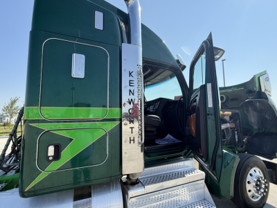 Kenworth W990 truck cab with open door, visible side step and vertical 'KENWORTH' lettering on the fuel/tank guard. The door area shows part of the cabin interior and steps with metal grating.