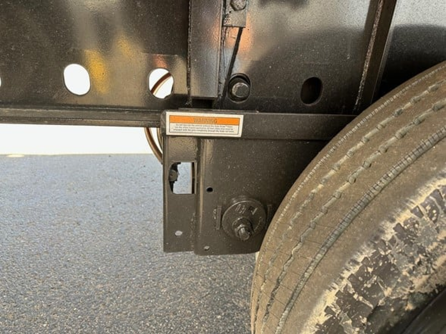 Trailer frame with wheel and mounting bracket showing a small orange warning label and bolt, side view of a utility flatbed trailer hitch area.