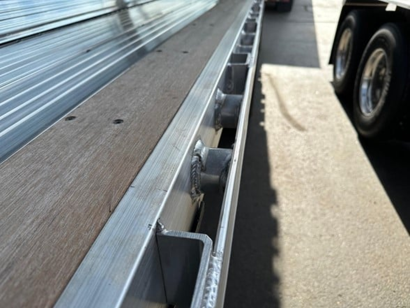 Flatbed trailer edge with wooden decking and metal side rails, close-up view showing stake pockets and reflective hardware.