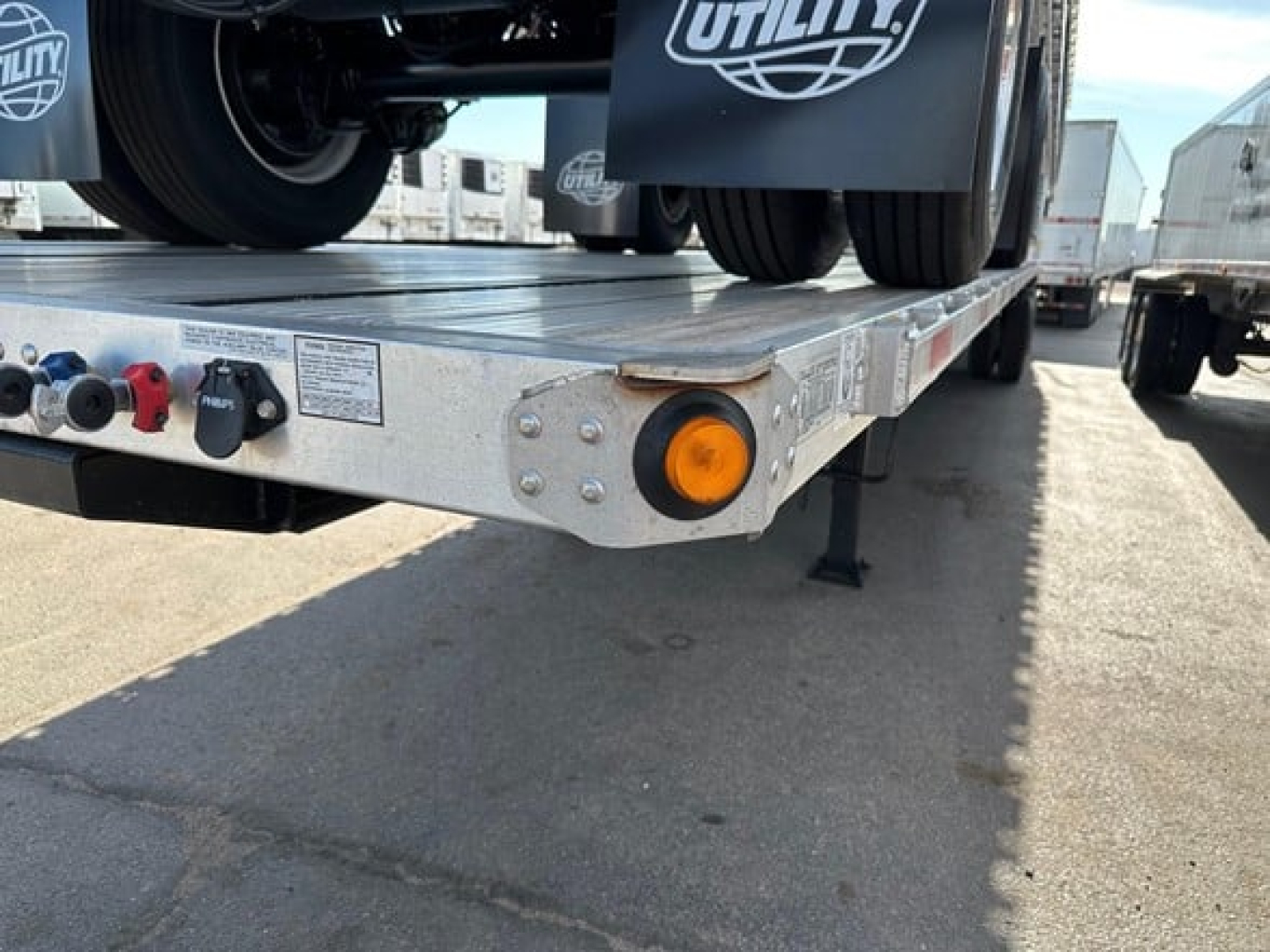 Utility flatbed trailer rear-left corner showing amber side marker light, power-outlet panel, and bumper with black wheels in background.