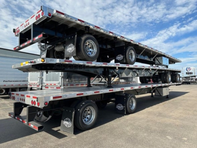 Flatbed trailer stack showing multiple gold trailers with wheels, tires, and brand decals. Visible text includes company logos on mudflaps and reflective safety markings along edges.