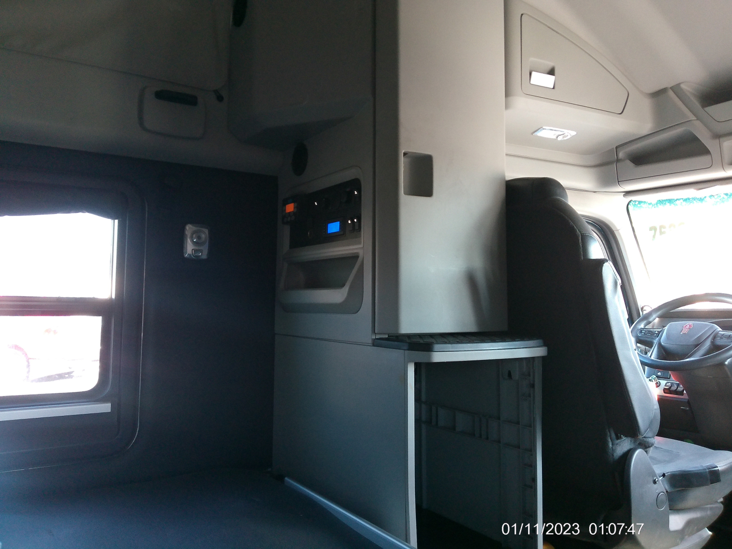 Cab interior of a Kenworth tractor showing storage cabinet with a small computer or radio panel, overhead lighting, and driver's seat; window on left.