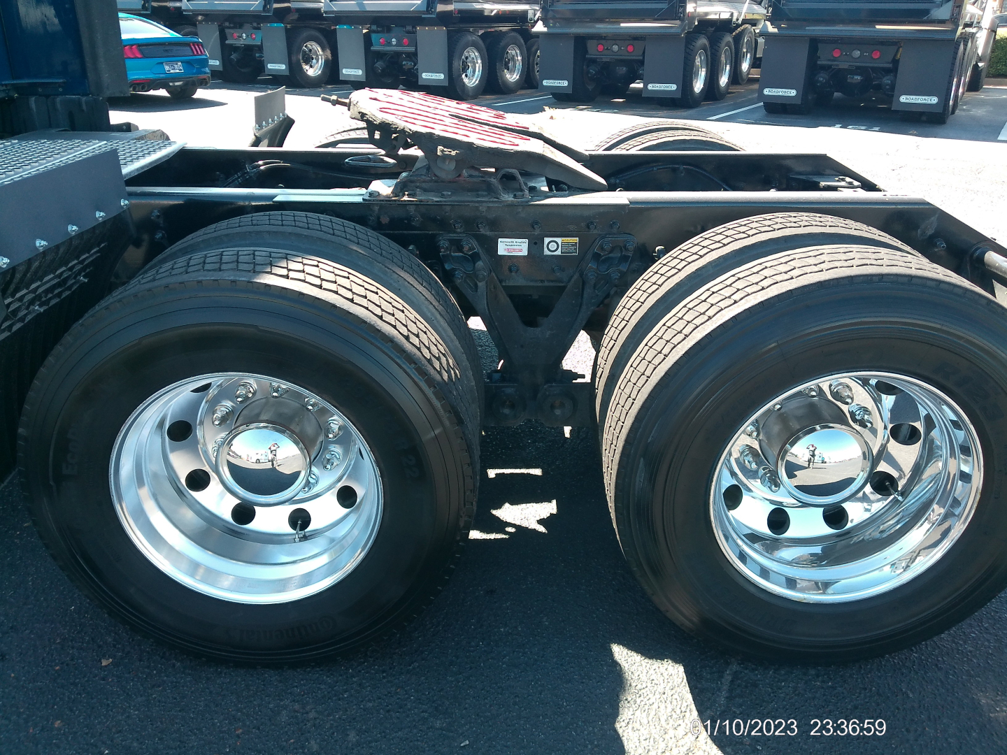 Kenworth T680 rear wheels and suspension showing dual rear axle with chrome rims and tires; photo includes undercarriage and adjacent trucks in lot.