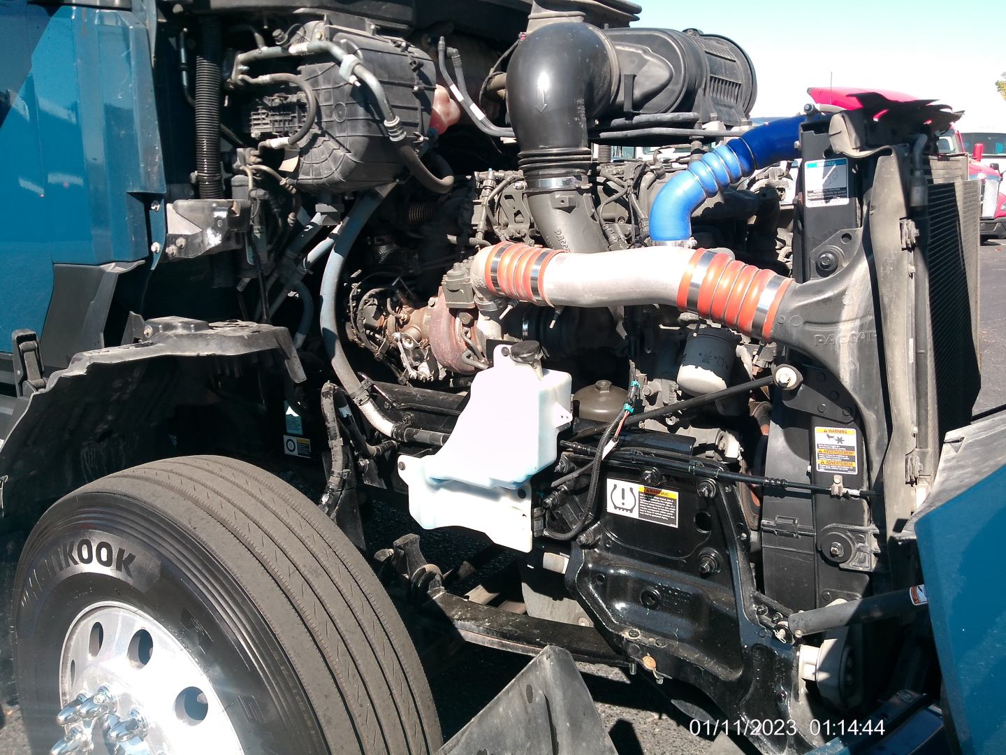 Kenworth T680 engine bay with visible intake piping, hoses, and radiator area; front-left wheel visible