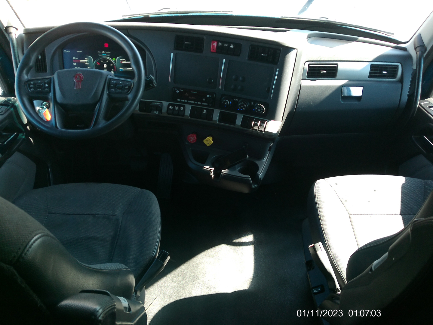 Kenworth T680 dashboard and driver seat view showing steering wheel with red emblem, instrument cluster, center console, and passenger seat in a cab interior.