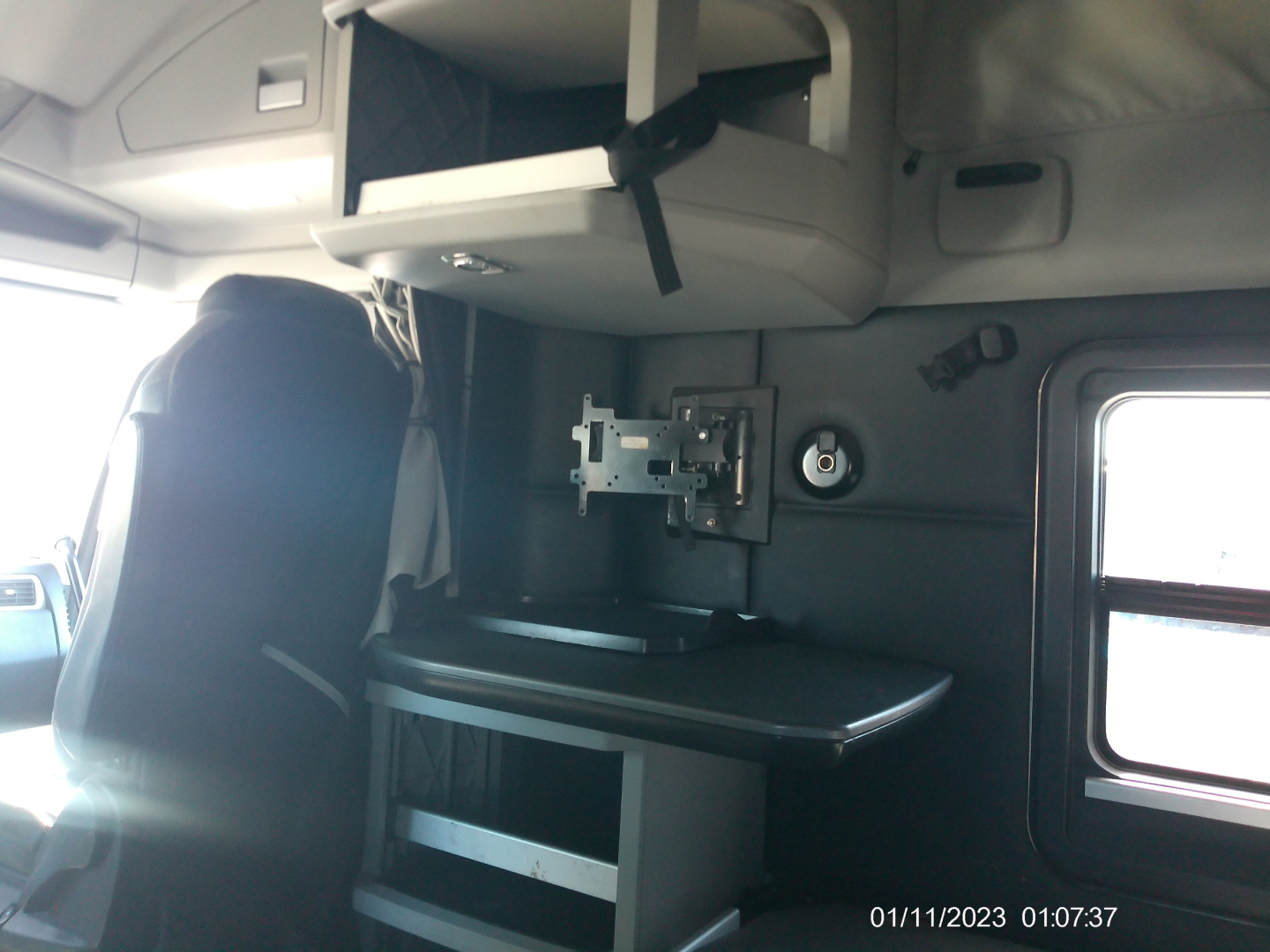 Trucks interior showing sleeper cab with mounted TV monitor, shelf, and workstation area. Passenger seat and window visible.