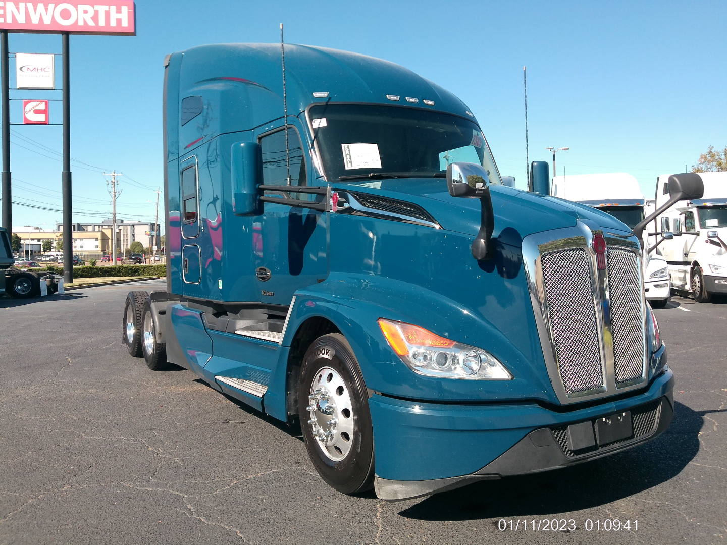 Kenworth T680 semi-truck in blue paint, side view showing front grille, headlights, mirrors, and cab with sleeper area; stock lot in background.