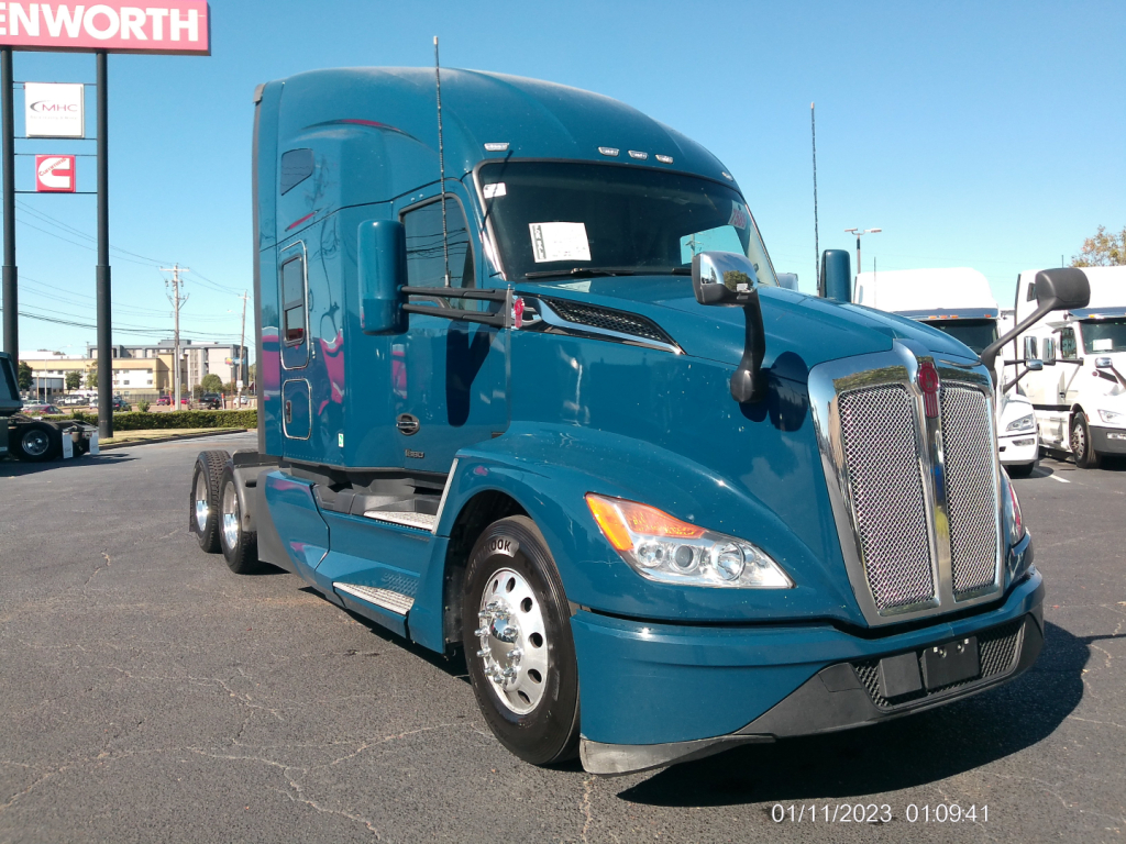 Kenworth T680 semi-truck in blue paint, side view showing front grille, headlights, mirrors, and cab with sleeper area; stock lot in background.