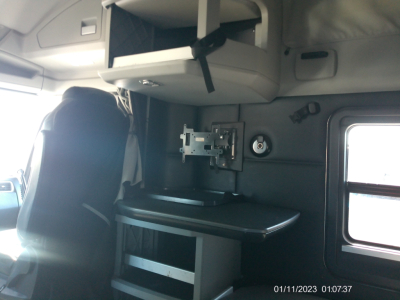 Trucks interior showing sleeper cab with mounted TV monitor, shelf, and workstation area. Passenger seat and window visible.