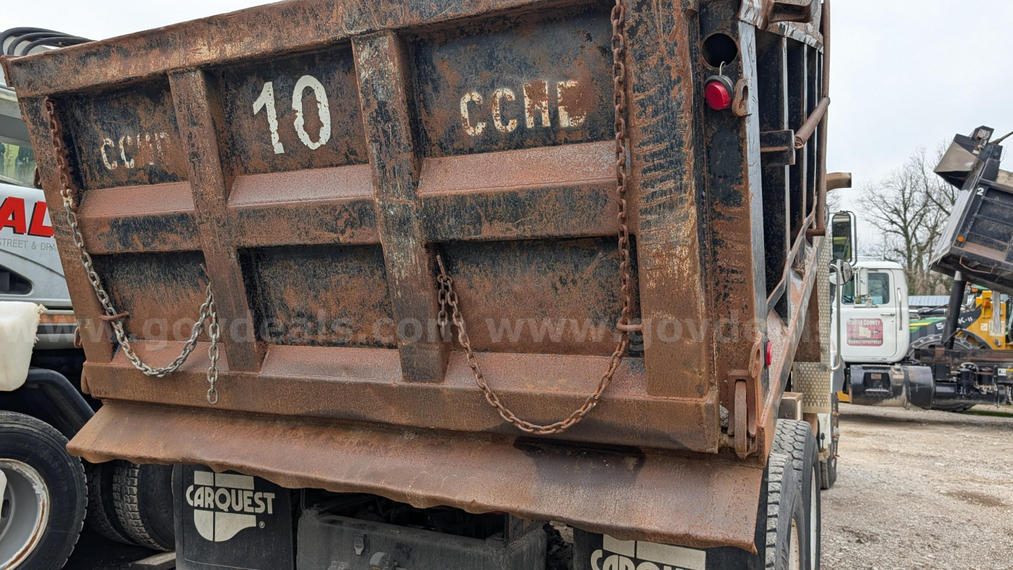 Dump truck bed showing rusted rear with 'CARHEST' markings, tailgate down, and chain latches; heavy-duty chassis and rear wheel visible.