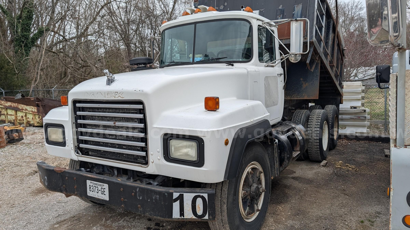 Dump truck cab and chassis showing white Mack RD690S with front grille, headlights, and large dump bed in background.