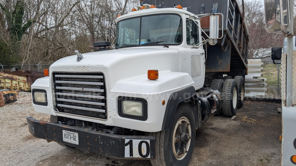 Dump truck cab and chassis showing white Mack RD690S with front grille, headlights, and large dump bed in background.