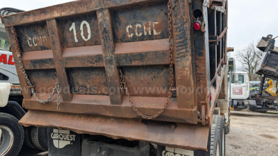 Dump truck bed showing rusted rear with 'CARHEST' markings, tailgate down, and chain latches; heavy-duty chassis and rear wheel visible.