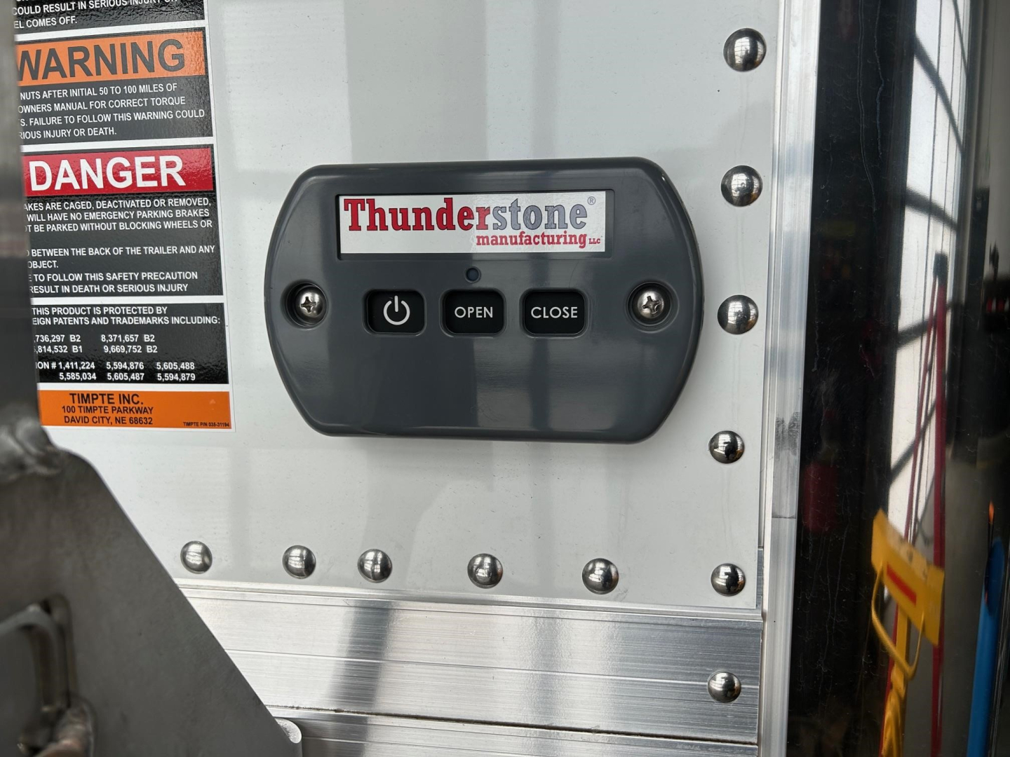 Close-up of a Thunderstone Manufacturing control panel on a Timpte hopper trailer; panel has OPEN and CLOSE buttons and a Thunderstone logo; warning labels on left edge