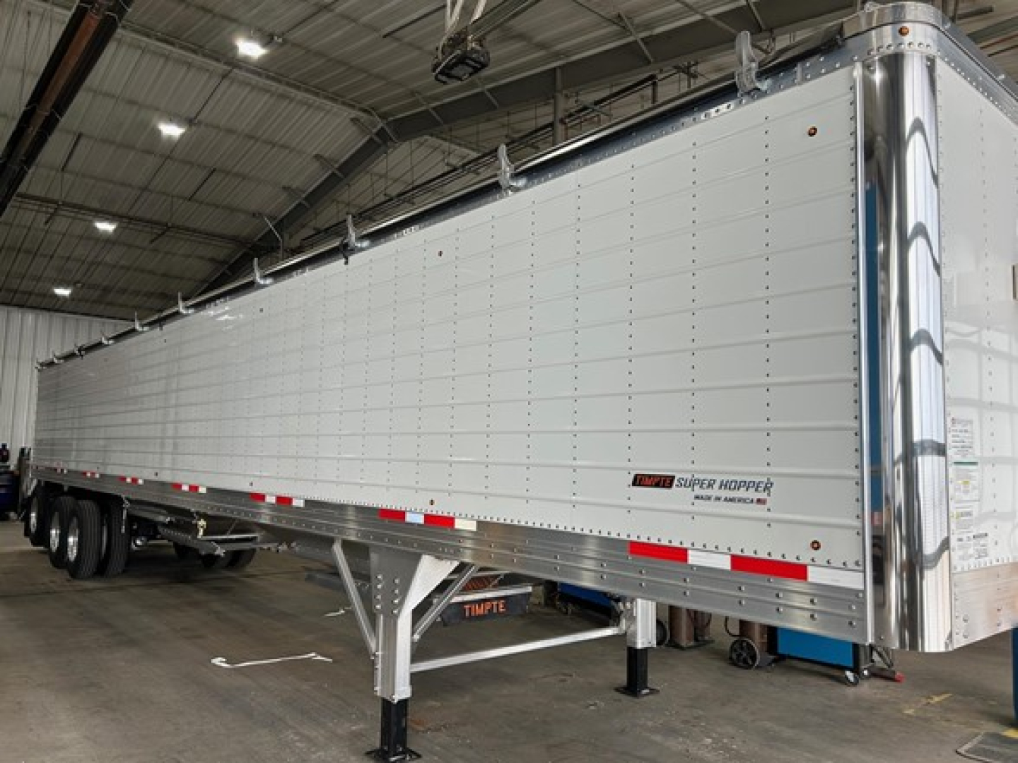 Timpte Super Hopper trailer in a showroom, long aluminum side panels with reflective red/white rails, rear doors closed, dual rear axles, landing gear visible.