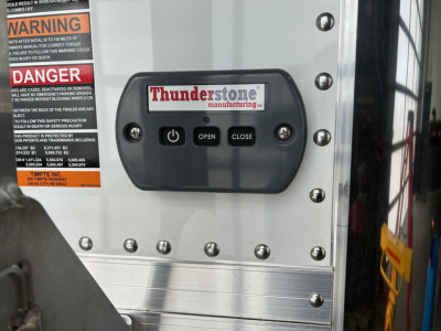 Close-up of a Thunderstone Manufacturing control panel on a Timpte hopper trailer; panel has OPEN and CLOSE buttons and a Thunderstone logo; warning labels on left edge
