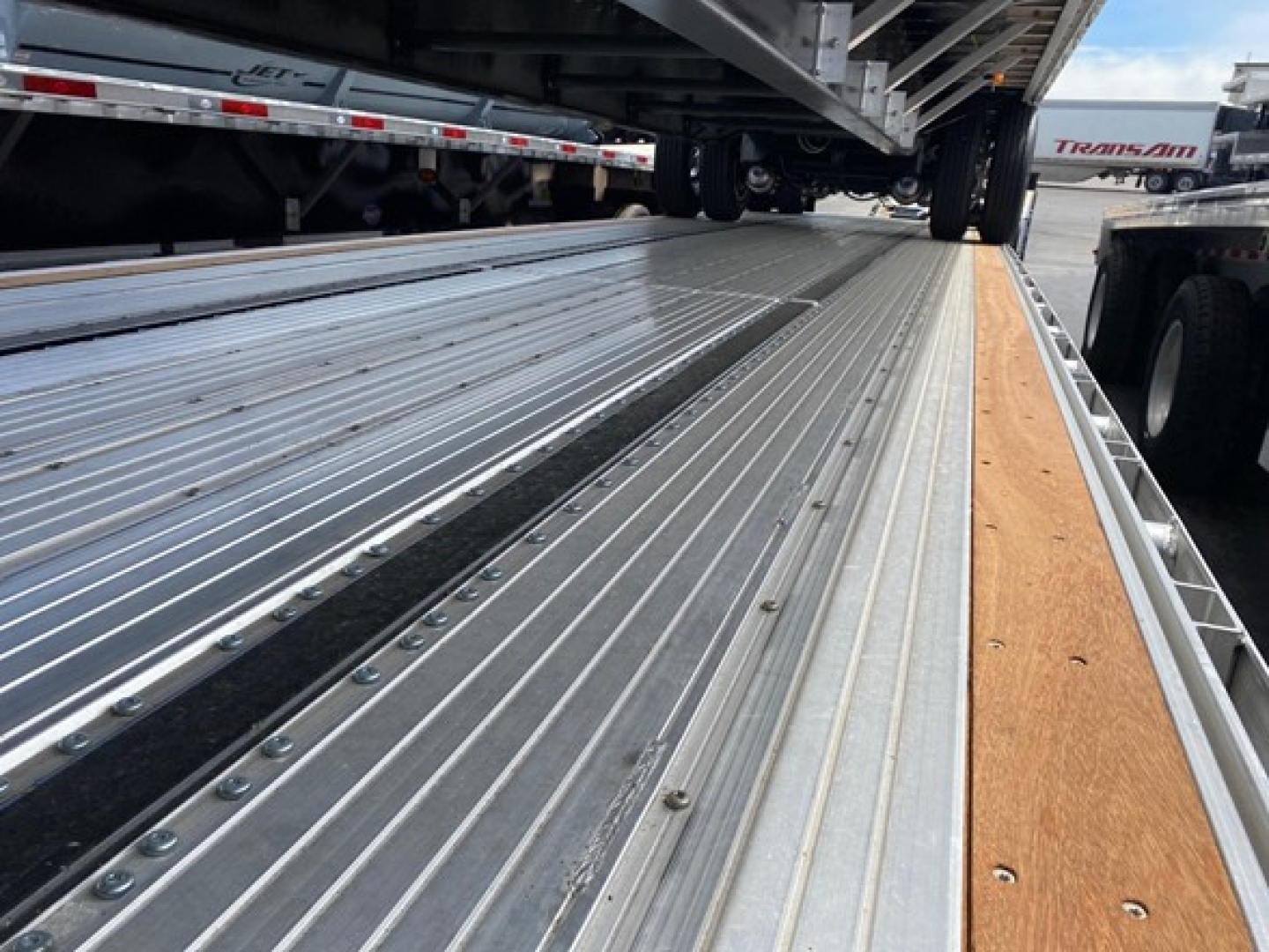 Trailer deck edge with metal slats and wooden side strip, perspective along length of a drop deck trailer showing riveted flooring and wheels in background.