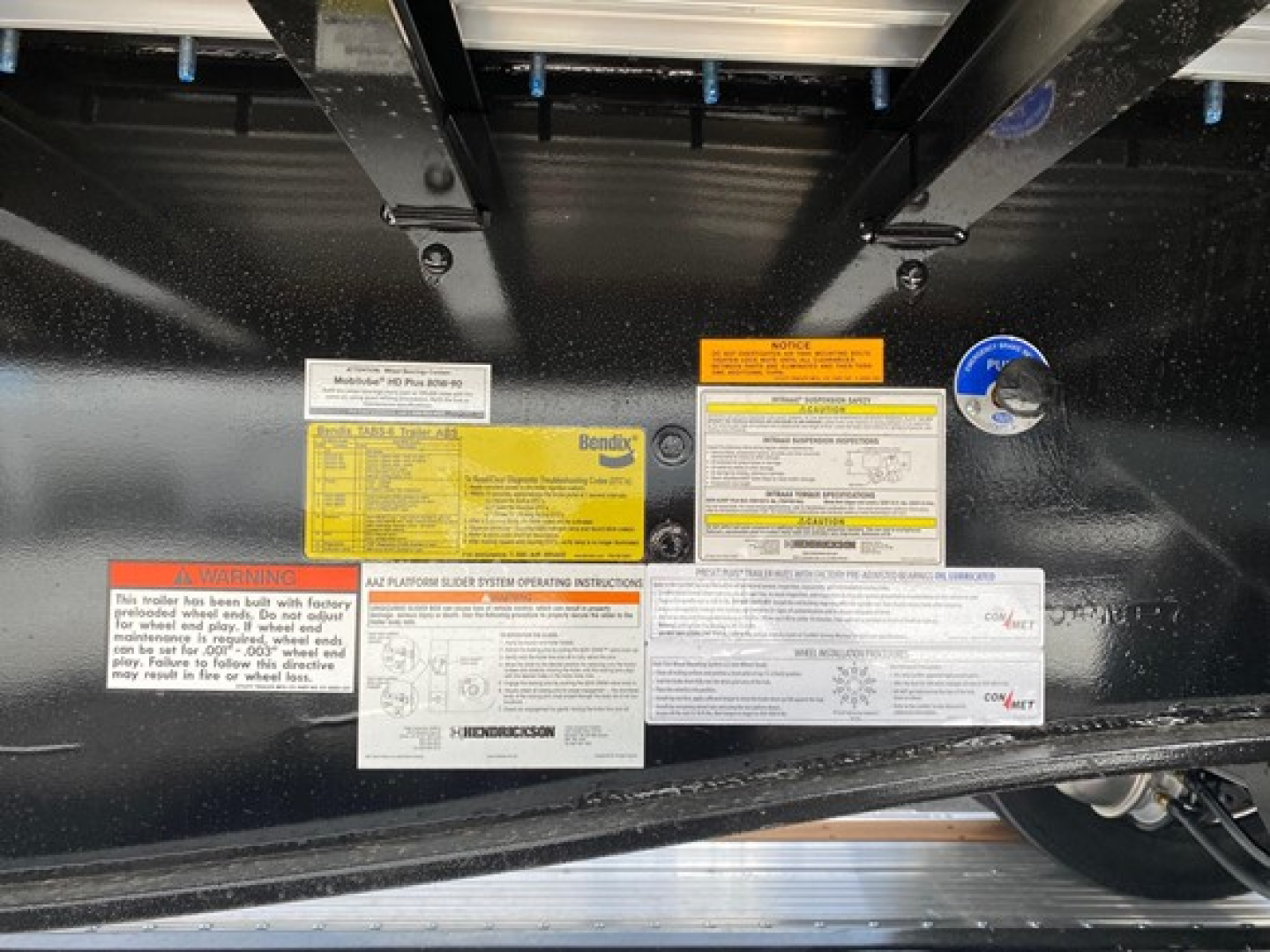 Trailer deck underside showing multiple instruction and warning labels on black metal frame; includes yellow, white and orange labels with text, and a blue circular sticker.