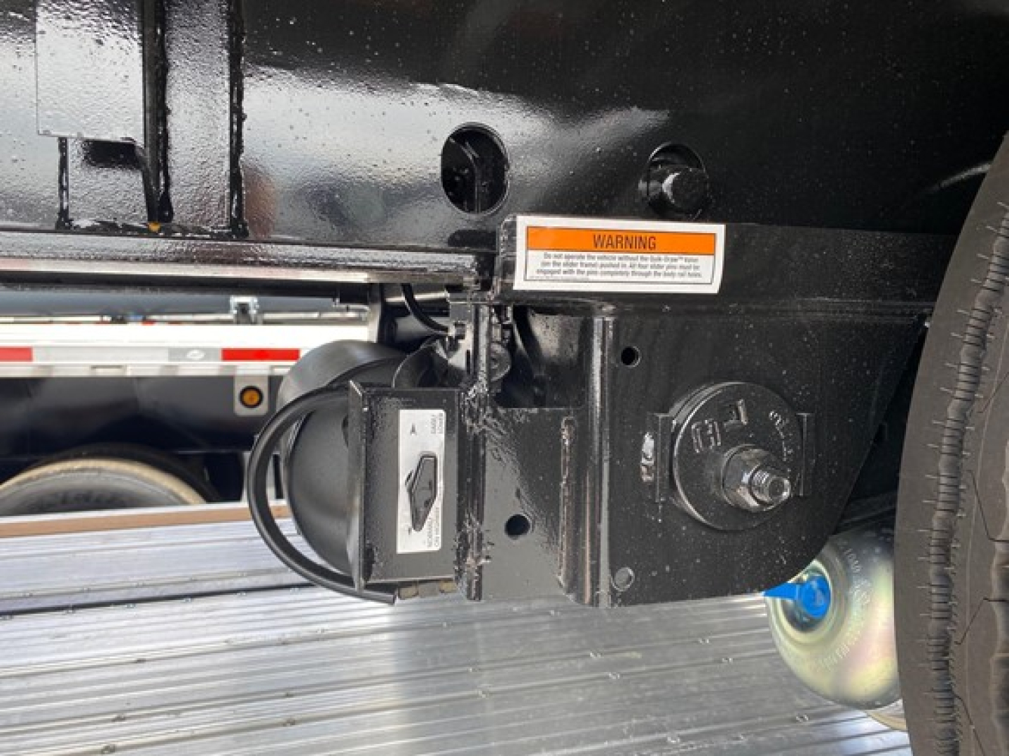 Utility trailer hitch area showing a circular 7-pin connector and a small electric control plug, warning sticker, and steel frame.