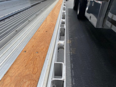 Trailer deck with wooden center plank and metal side rails showing tie-down slots and a parked truck on the right.