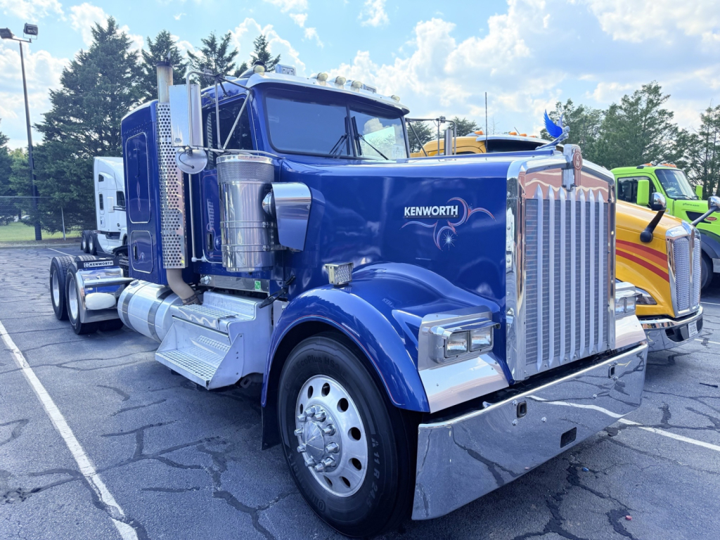 Kenworth W900L blue semi-truck front-left view showing chrome grille, large vertical exhaust stacks, side fuel tanks, and dual rear wheels.