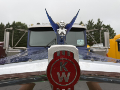 Kenworth W900L truck front grille emblem with KW letters and hood ornament; windshield and trucks in background; no license plate visible.