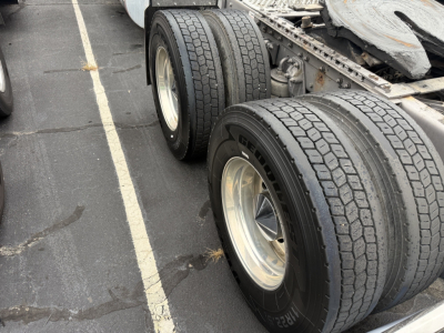 Kenworth W900L truck rear dual rear wheels and chassis shown in parking lot; visible tires and rims; partial trailer frame and mudguard edge.