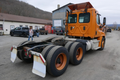 Orange Freightliner Cascadia 125 day cab tractor with dual rear axles and fifth wheel, seen from rear-left, VIN 1FUJGEDV5HLHZ1375 on lot.