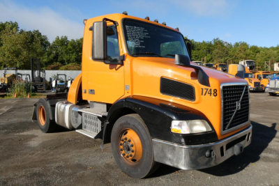 Volvo VNM42T conventional daycab truck in orange with black lower front, showing 1748 on door and VIN/stock text on window.