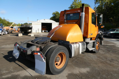 Volvo VNM42T conventional daycab tractor in orange with rear coupler and dual rear wheels, side exhaust, cab windows, and engine bay area visible.