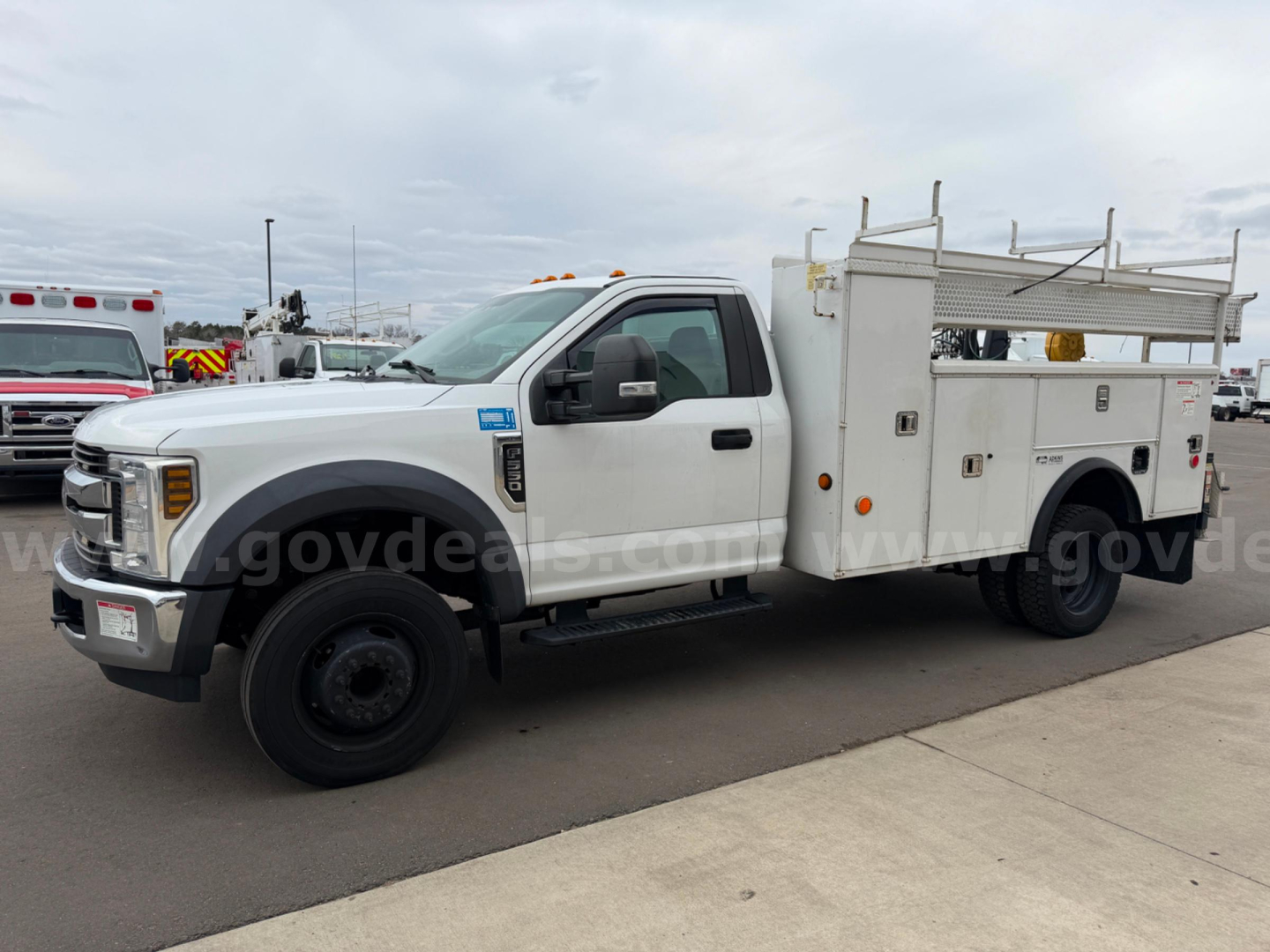 White Ford F-550 service utility truck with utility body and ladder rack, side view showing storage compartments and front grille.
