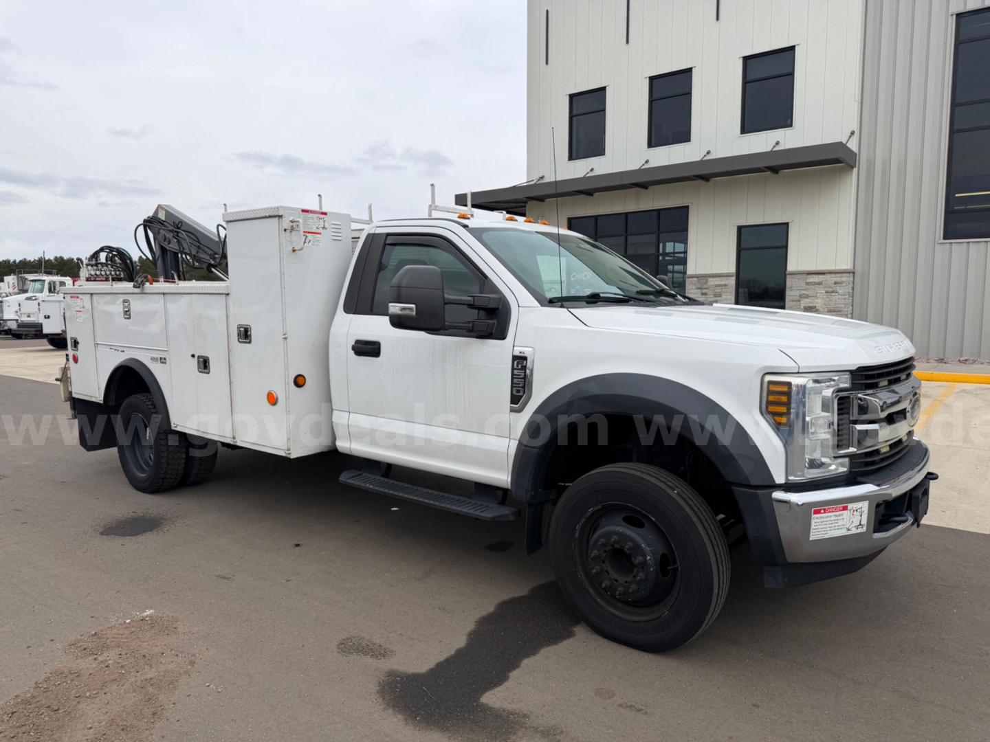 Service/utility Ford F-550 truck side view showing utility bed with storage compartments and crane mount. White cab with black fenders, stock number plate visible.