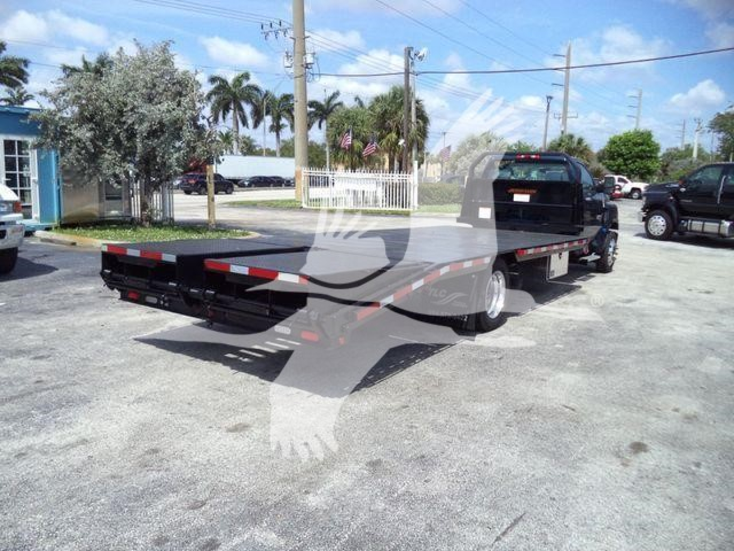 Flatbed tow truck trailer attached to a Silverado 6500HD chassis in a parking lot. Side view shows long flatbed with low-profile wheel wells and rear ramps; clear blue sky.