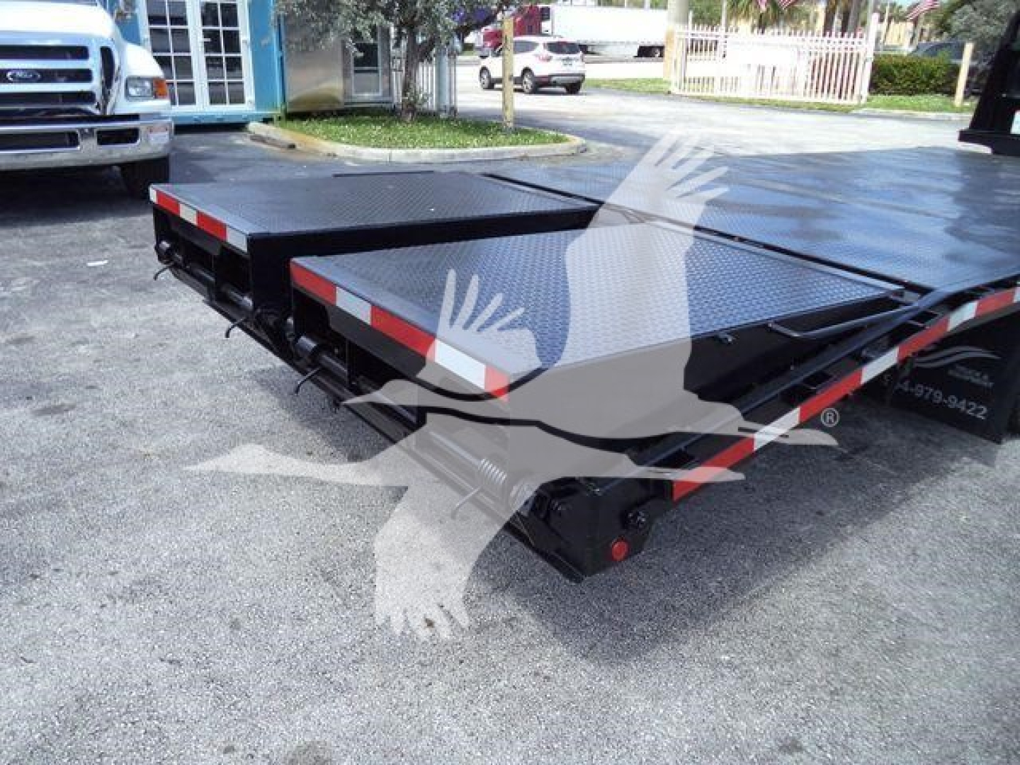 Flatbed trailer deck with black diamond-plate surface and red-white reflective tape, viewed from rear corner.
