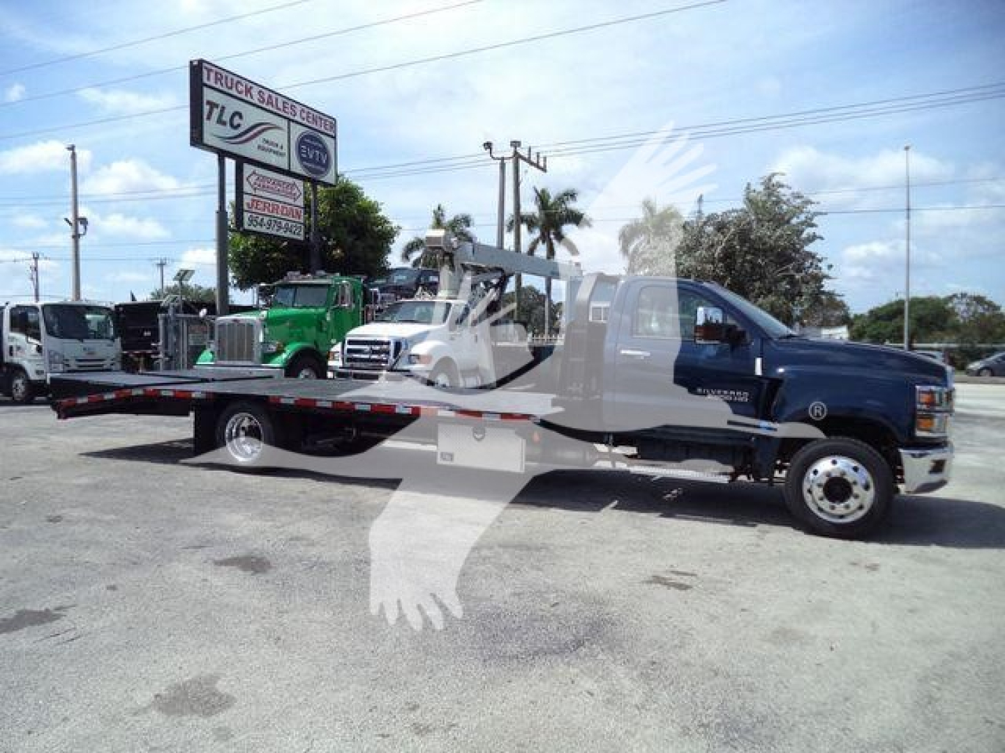 Chevrolet Silverado 6500HD tow truck on a flatbed trailer, blue cab and dark chassis, dealership lot in background.
