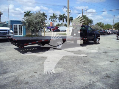 Flatbed tow truck with black cab on a dealership lot; faint white vehicle silhouette watermark overlay. Visible text: none legible on signs due to overlay.
