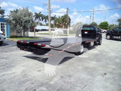 Flatbed tow truck trailer attached to a Silverado 6500HD chassis in a parking lot. Side view shows long flatbed with low-profile wheel wells and rear ramps; clear blue sky.