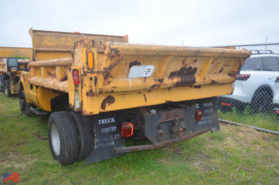 Dump truck bed of a yellow 2009 Ford F350 Super Duty dump truck showing tailgate area with black metal mudflaps and faded paint. Visible text includes 'TRUCK EQUIP' on tailgate and small serial plate, and a white label on bed side.