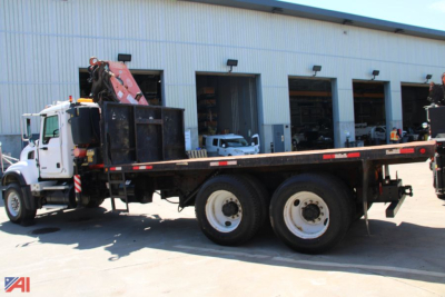 Tractor-trailer style flatbed truck with a hydraulic knuckle boom crane mounted on a long flatbed; white cab, dual rear axles, crane in folded position near cab; light-industrial building in background.
