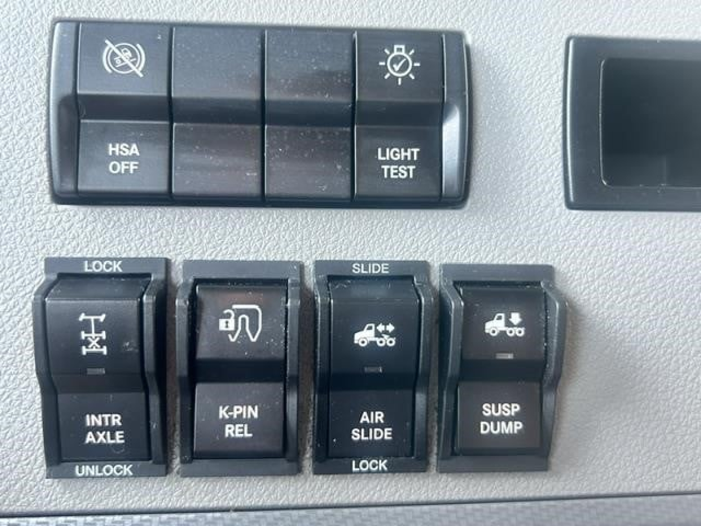 Panel of switches and controls in Freightliner Cascadia cab showing HSA OFF, light test, lock/slide, air slide, susp dump, and other labeled buttons.