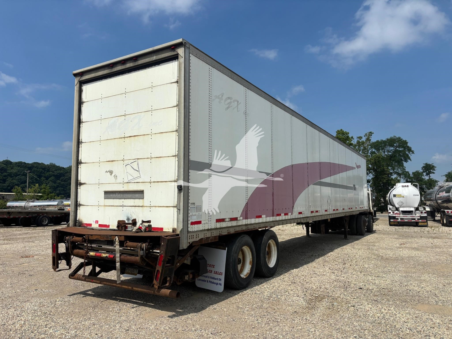 Rear three-quarter view of a Wabash dry van trailer with a purple bird graphic, showing rear doors, wheels, and undercarriage. Some rust marks on doors and frame, gravel lot in background.