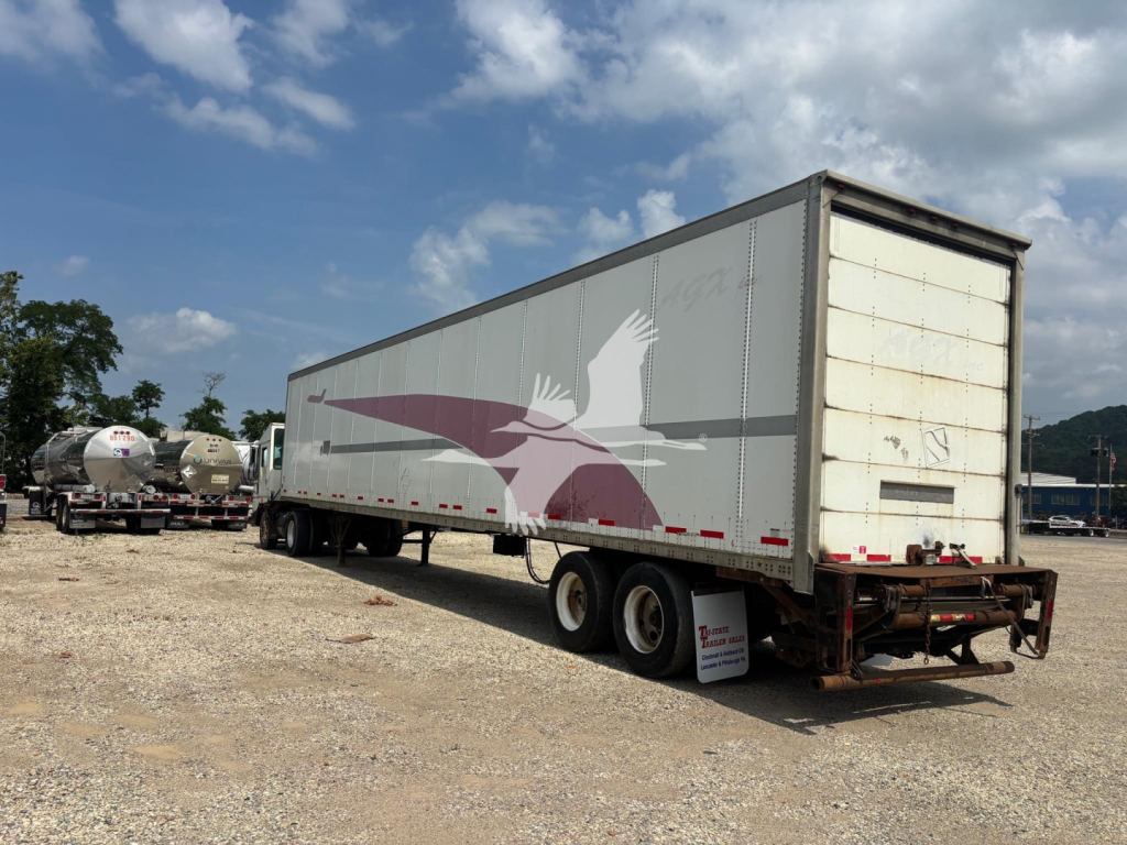Trailer side view showing a gray Wabash DVCVHPC trailer with a maroon and white eagle graphic, tandem axles, rear roll-up door, empty lot background.