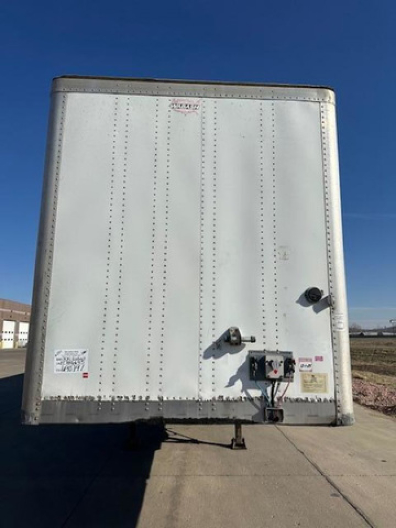 Trailer rear view showing white bonded metal van body, doors closed with hitch and electrical connections, rear bumper, and small decals.