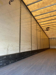 Trailer interior showing empty cargo van with wooden floor and side panels, warehouse lighting reflections, and ceiling ribs.