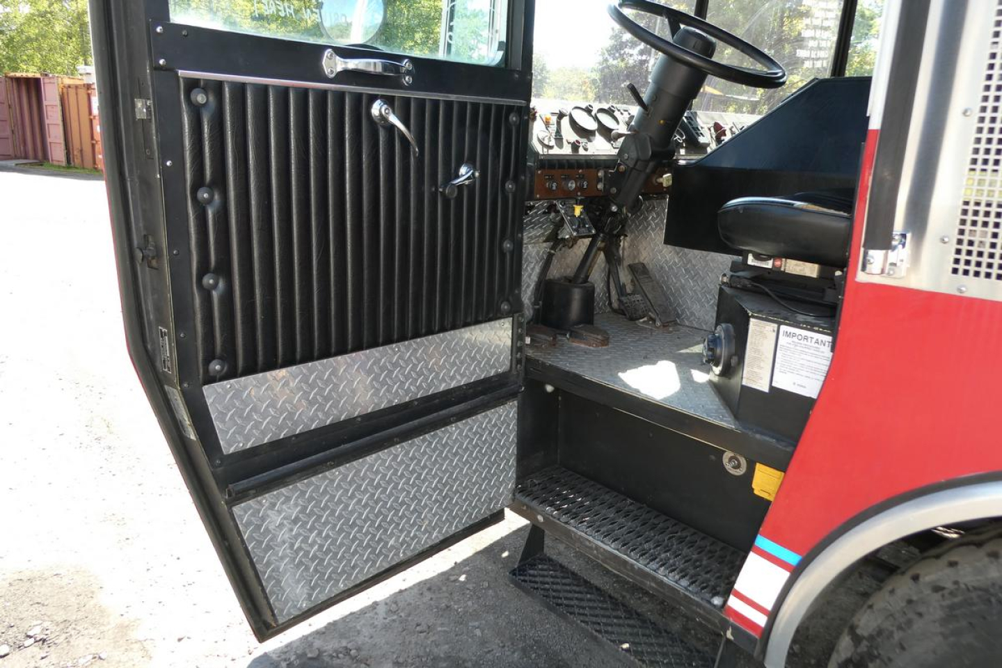 Fire truck cab interior with open door showing black ribbed door panel, metal diamond plate step, steering wheel, and dashboard controls.