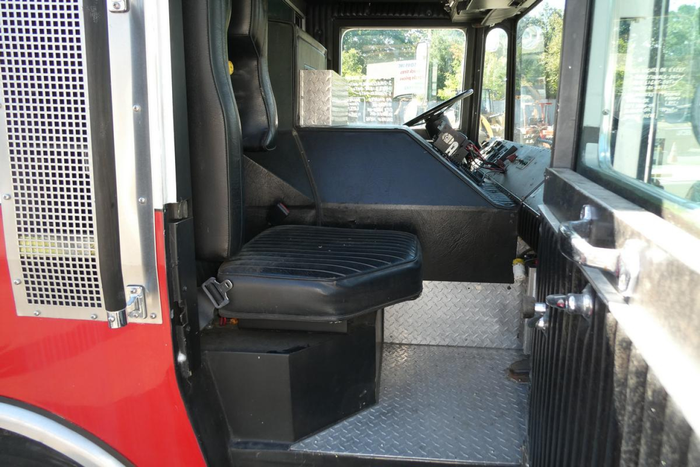 Fire truck cab interior showing driver seat and control dashboard, metal diamond-plate floor, and side window; red exterior visible at left.